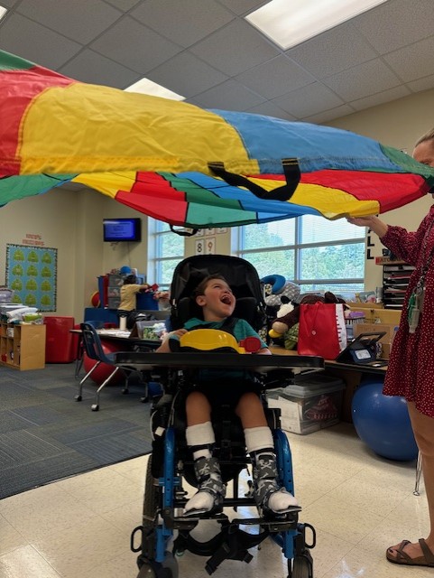 A young boy in a wheelchair smiles and looks up as a colorful parachute is lifted above him in a classroom. An adult holds the parachute, and classroom supplies are visible in the background, capturing a joyful moment from Jake's Story.