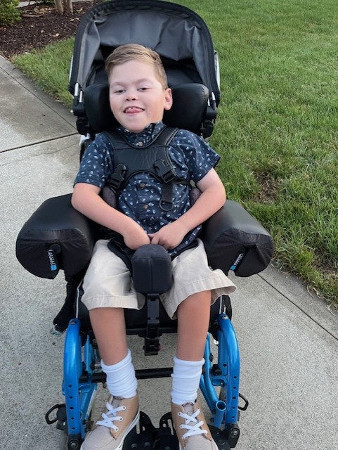 Jake's Story: A young boy with light brown hair sits in a blue adaptive wheelchair on a sidewalk. He is smiling, wearing a navy patterned shirt, tan shorts, white socks, and brown shoes. Green grass stretches in the background.