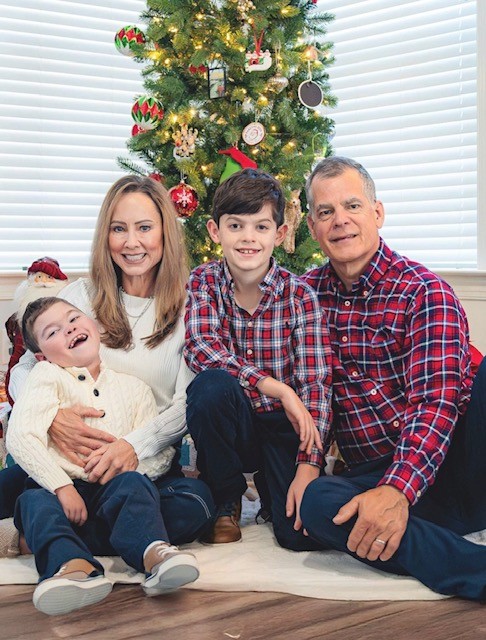 A family of four sits and smiles in front of a decorated Christmas tree. This could be Jake's Story—parents on either side, with their two young sons between them, all wearing festive outfits in red, white, and blue tones.
