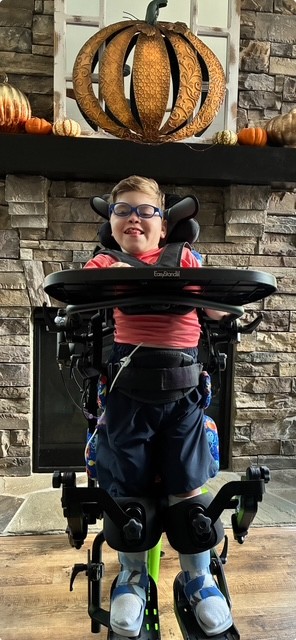 A young boy wearing glasses and a red shirt stands in a wheelchair in front of a stone fireplace decorated with pumpkins, with a large pumpkin ornament on the mantel—capturing a special moment from Jake's Story.