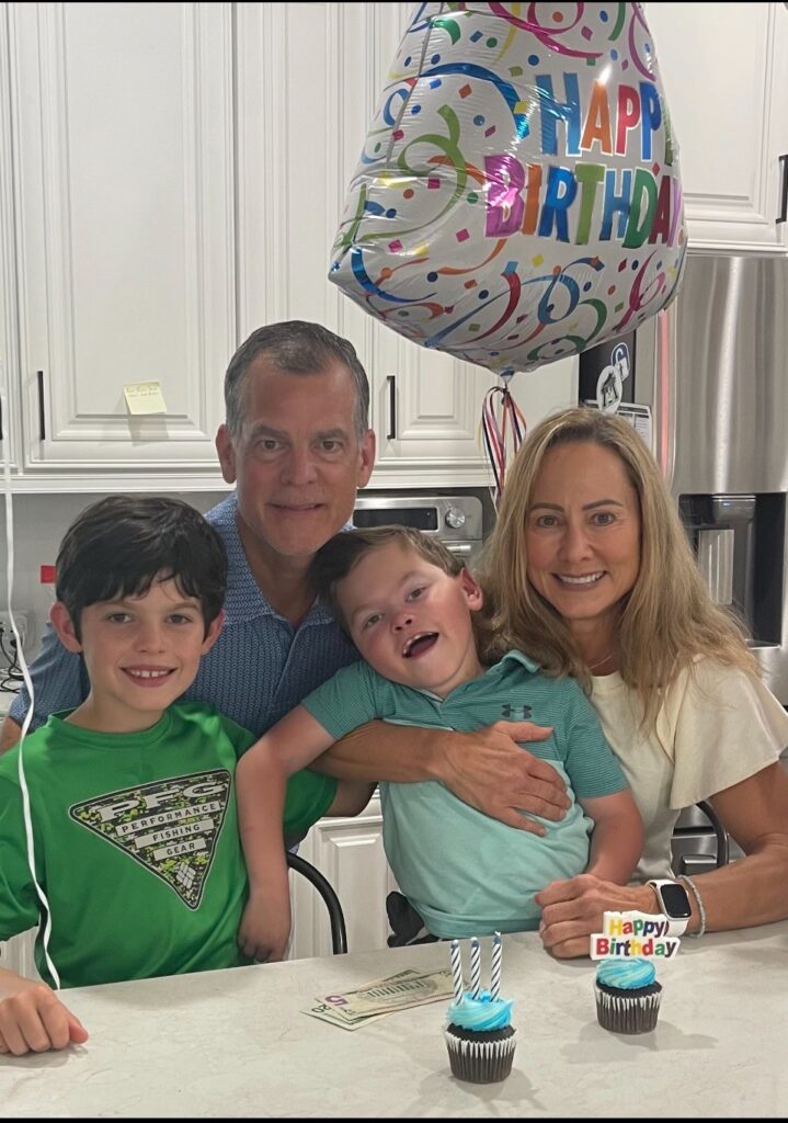 A family of four smiles at a kitchen counter with two birthday cupcakes and a Happy Birthday balloon. The parents sit behind their two young sons, celebrating Jake's Story in a bright, modern kitchen.