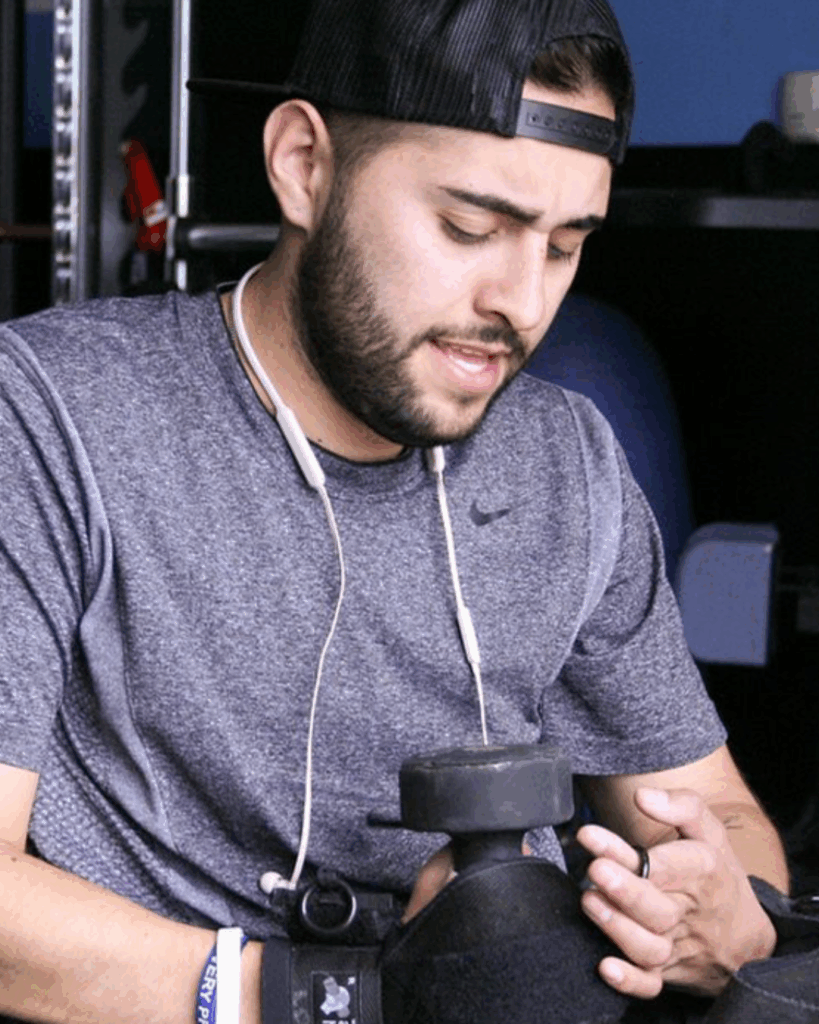 A man wearing a backwards cap, gray athletic shirt, and earphones looks down while holding a dumbbell with both hands in a gym setting.