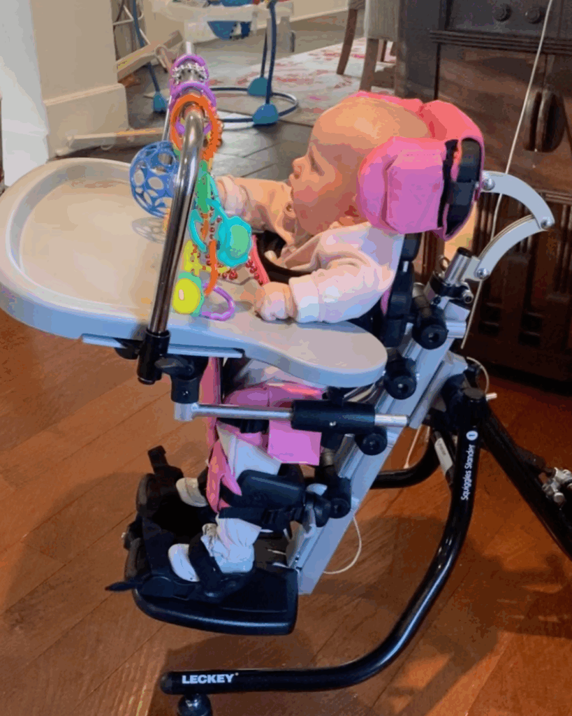 A baby in a pink outfit sits in a supportive adaptive chair, playing with colorful sensory toys attached to the tray. The scene, part of Abigail's Story, features black and pink padding and is set on a wooden floor indoors.