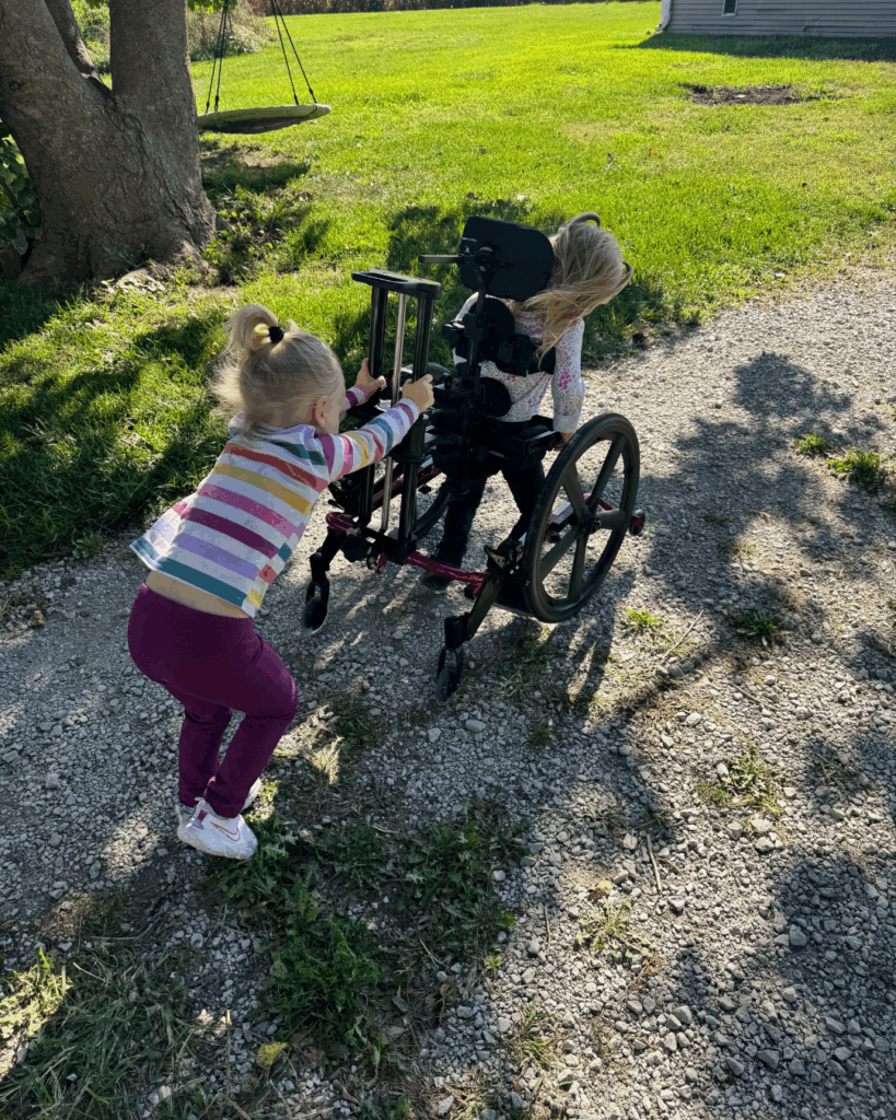 Two young girls are outside on a sunny day. In Abigail's Story, one girl in a wheelchair faces away, while her friend in a striped shirt and ponytail pushes her along a gravel path near green grass and trees.