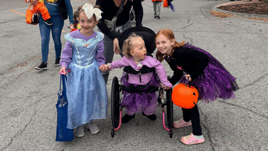 Three children in Halloween costumes smile outdoors. One wears a blue princess dress, another is in a wheelchair wearing a lavender outfit from Abigail's Story, and the third sports a black and purple tutu, holding a pumpkin bucket. An adult stands behind them.