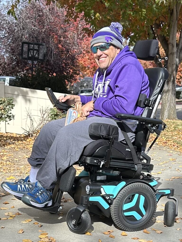 A smiling man wearing a purple hoodie, gray sweatpants, blue sneakers, and a beanie sits in a turquoise power wheelchair on a sunny autumn day, surrounded by colorful fall leaves—capturing a moment from David's Story.