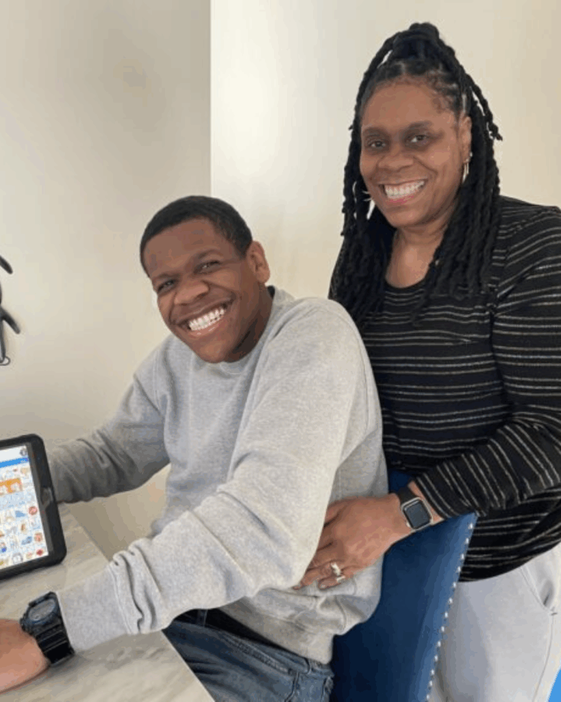 A young man sits at a table smiling broadly with a tablet in front of him, while an older woman stands beside him, also smiling, with her hand resting on his back. Both appear happy and relaxed indoors.