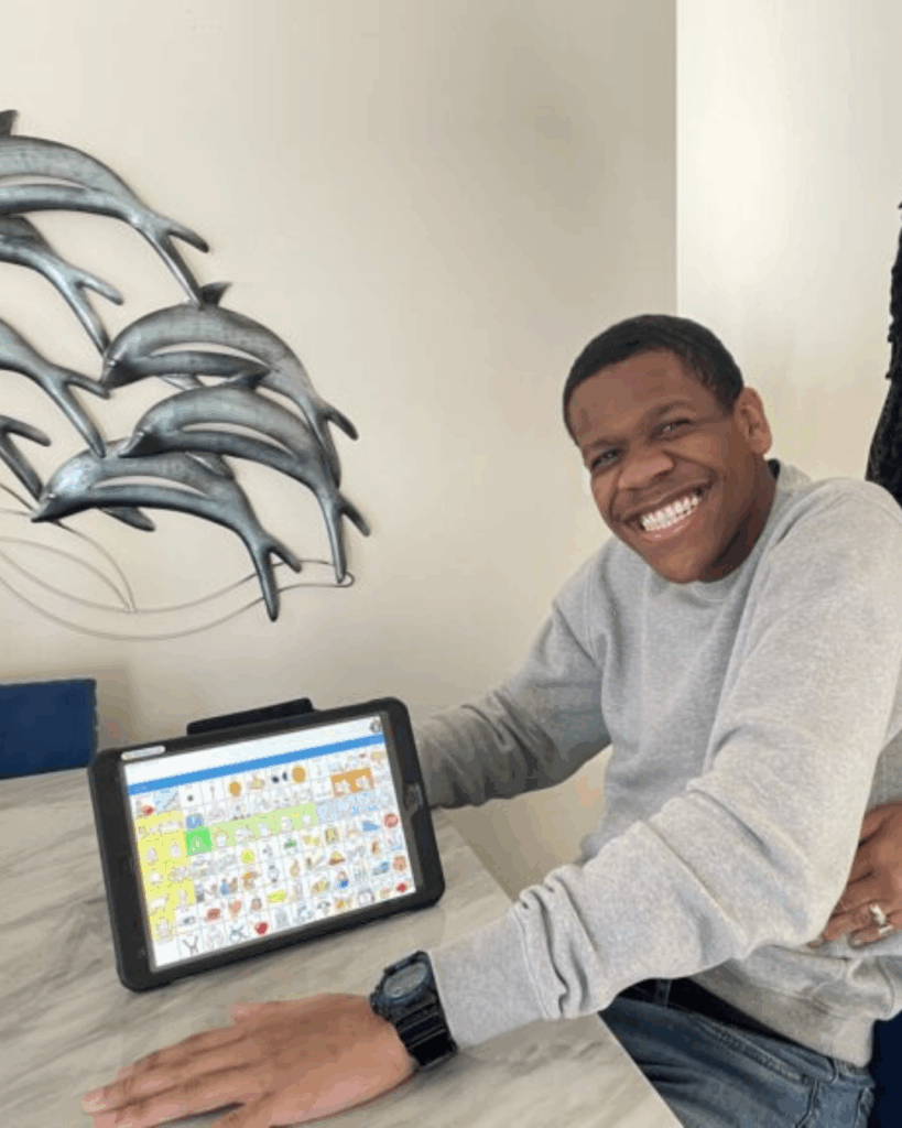 A smiling young man in a grey sweatshirt sits at a table, using a tablet with an augmentative and alternative communication (AAC) app. Metal dolphin wall art is visible in the background.