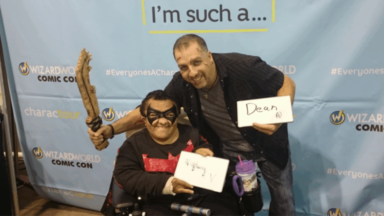 Two men pose and smile at a Comic Con event, sharing Zachary's Story. One is in a wheelchair, wearing a mask and holding a prop sword, while the other stands behind him with a "Dean" sign against a Wizard World Comic Con backdrop.