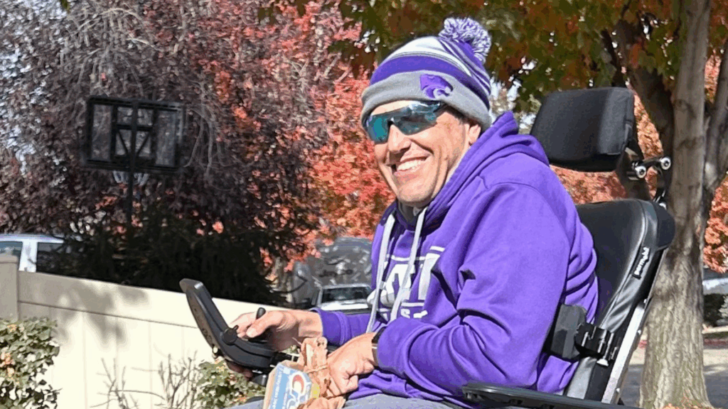 A smiling man in a wheelchair wears a purple hoodie, matching beanie, and sunglasses. He is outdoors with autumn trees in the background, enjoying a sunny day—another joyful moment from David's Story.