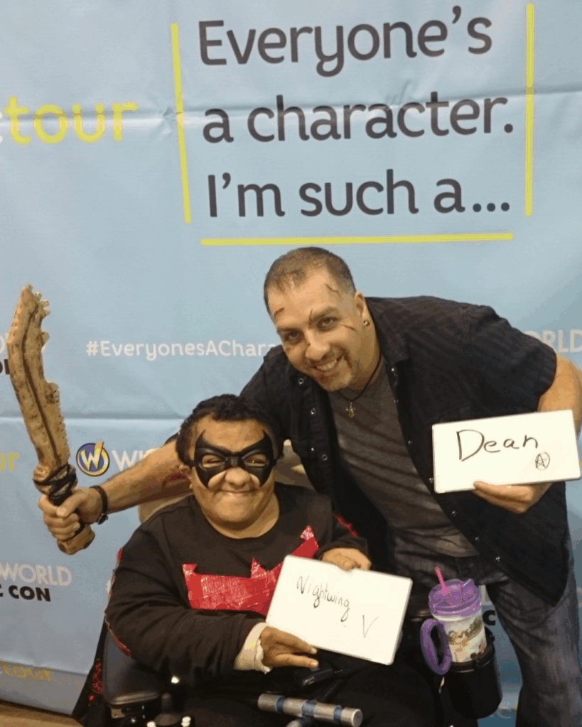 Two people pose in front of a banner reading Everyone’s a character. I’m such a…. One, in a wheelchair and costume with a mask, holds a sign reading Nightwing V—part of Zachary's Story. The other stands, smiling, holding a sign reading Dean.