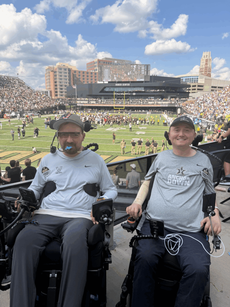 Two men in power wheelchairs smile at a football stadium, proudly wearing Vanderbilt gear. This moment, part of Josh's Advocacy Story, captures them in the stands with the field and city buildings bright on a sunny day.