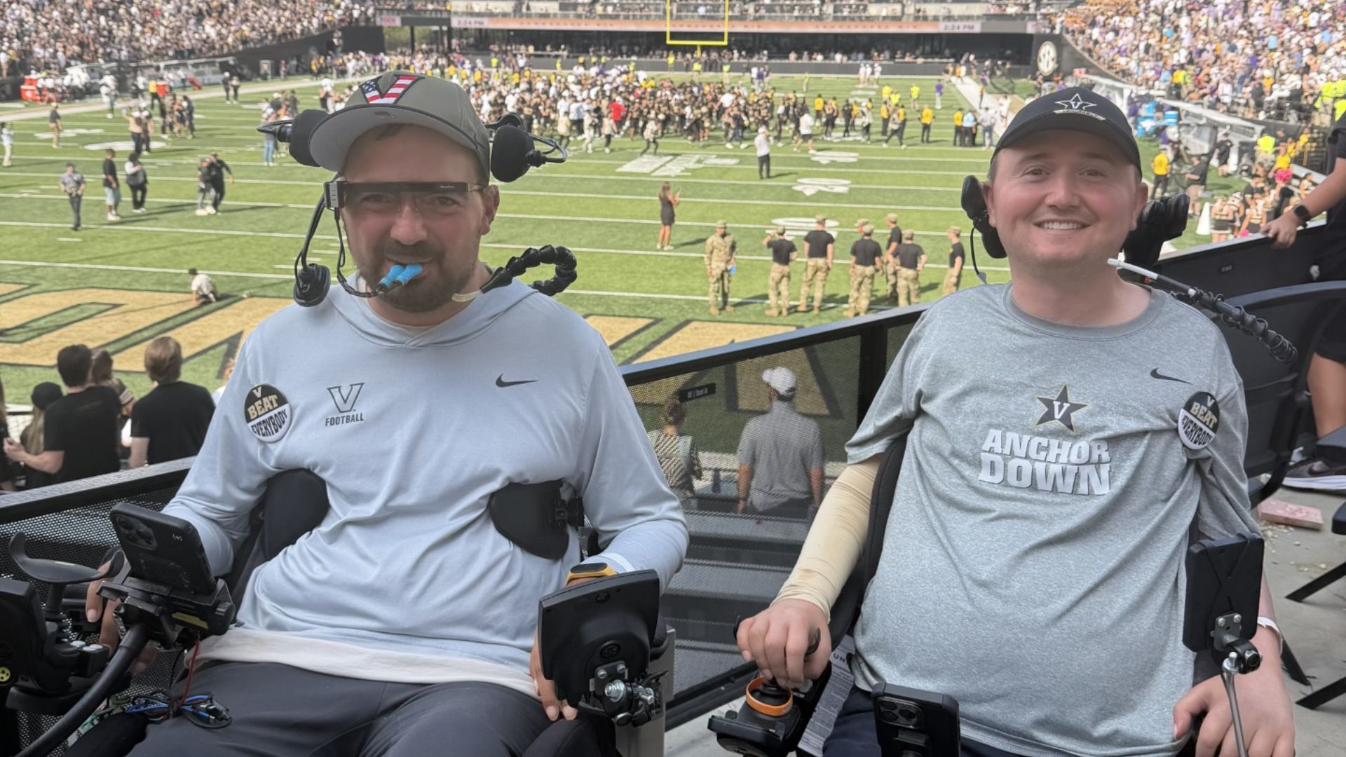 Two men in wheelchairs, both wearing Vanderbilt sports attire and caps, smile at the camera in a stadium filled with cheering fans—capturing a moment from Josh's Advocacy Story during an exciting football game.