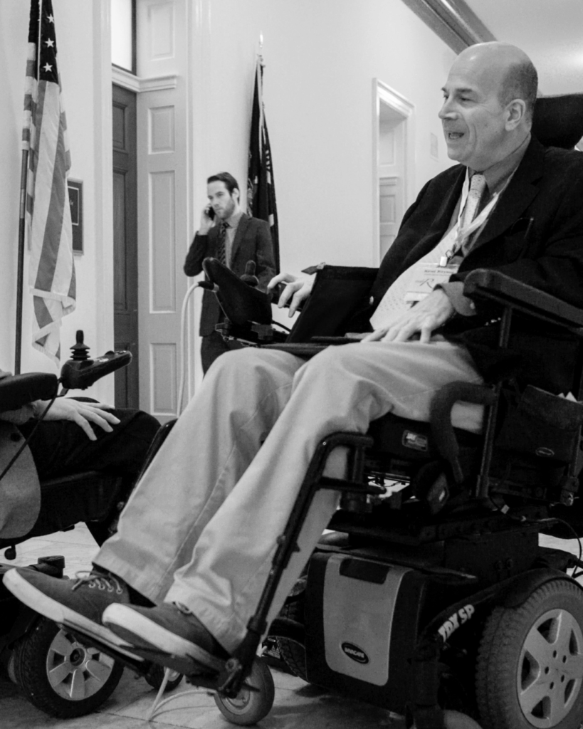 Two men in powered wheelchairs face each other in a hallway, embodying advocacy as they converse. One smiles, while another person in the background is on the phone. An American flag stands by an open doorway. The scene is in black and white.