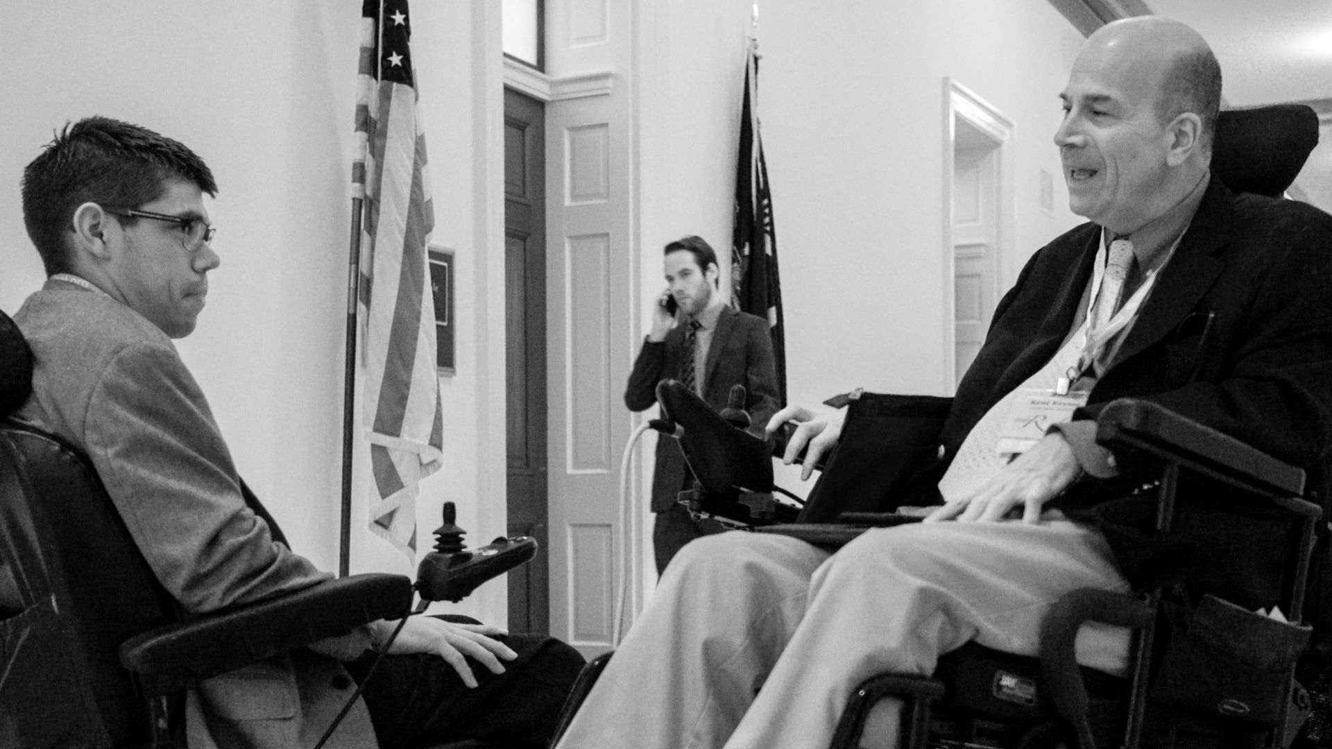 Two men in power wheelchairs engage in conversation in a hallway, highlighting Kent's Advocacy. An American flag and a man on the phone are visible in the background, suggesting this advocacy story unfolds inside a public or government building.