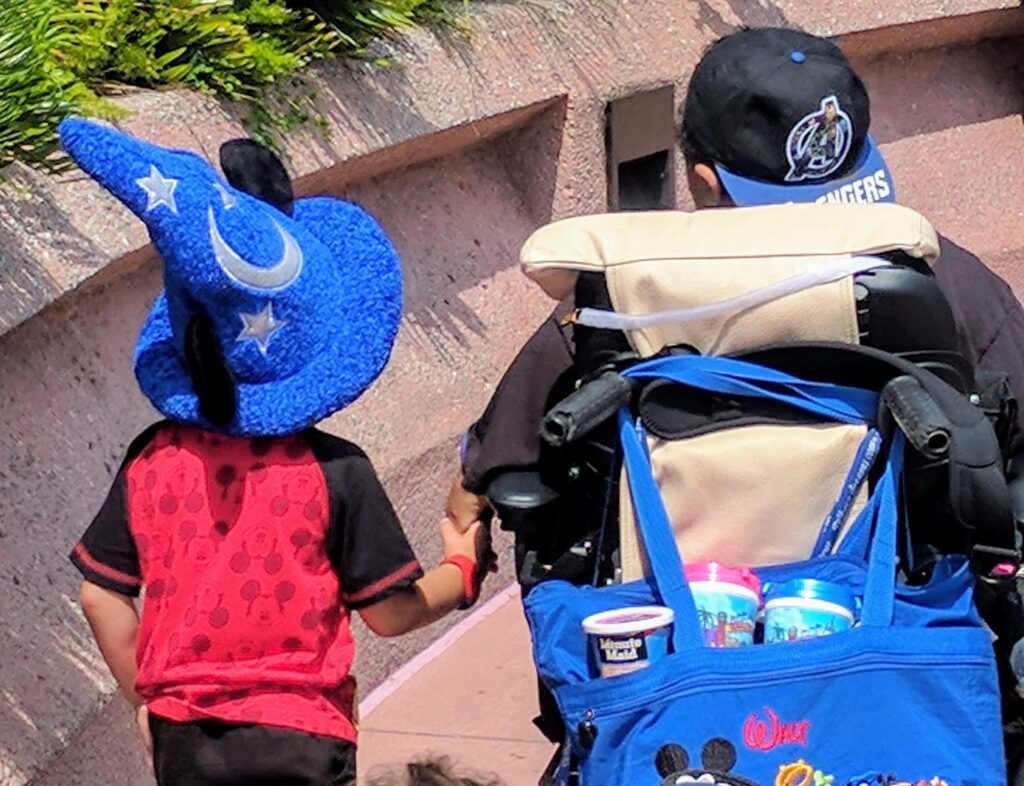 A child in a blue wizard hat with stars and a crescent moon holds hands with an adult in a wheelchair. The adult, wearing an Avengers cap, has a blue bag with drinks—capturing a magical moment from Zachary's Story.