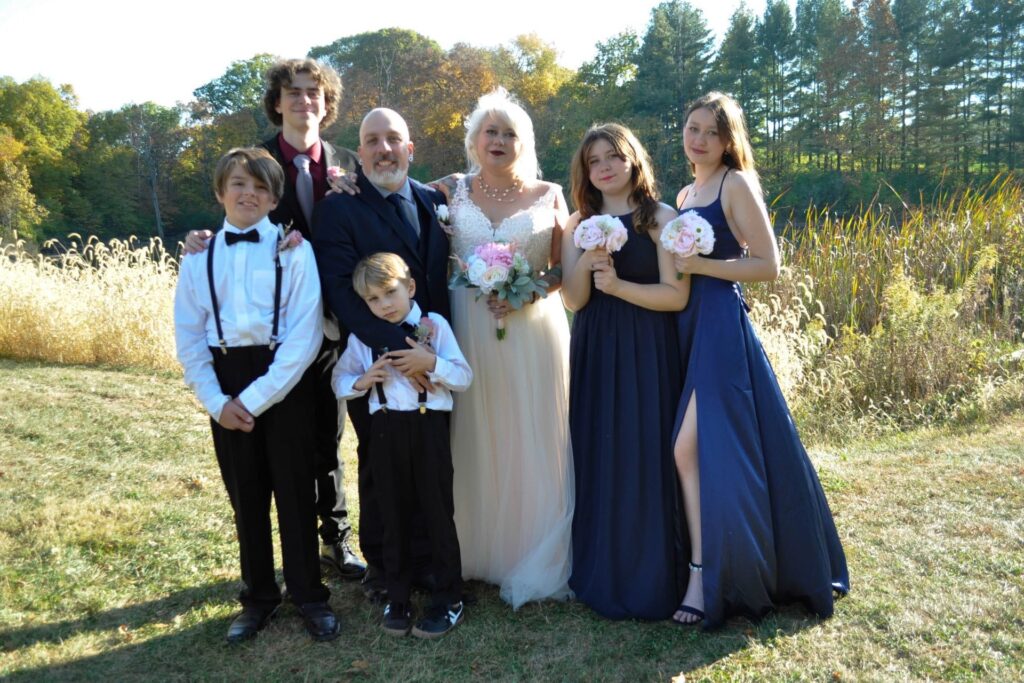 A bride and groom stand outdoors with four children and one adult, all dressed in formal attire. The group smiles at the camera, surrounded by tall grass and trees in bright daylight—capturing a joyful moment from Zachary's Story.