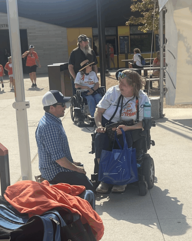 A man kneels to talk with a woman in a motorized wheelchair, who is holding a blue bag. Joey's Story unfolds as another woman in a wheelchair and a man stand behind them, while several people walk in the sunny background.