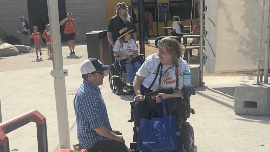 A man kneels and talks to a woman in a motorized wheelchair holding a blue bag. Another woman in a wheelchair and several people walk by, bringing Joey's Story to life in an outdoor public space on a sunny day.