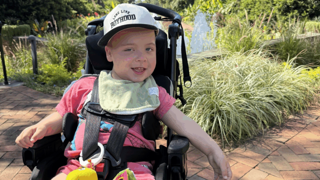 A young child in a wheelchair smiles outdoors on a sunny day, wearing a cap and pink shirt with a green bib. Lush greenery and a brick path set the scene for Myles' Story of joy and resilience.