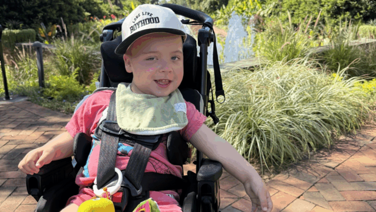 A young child in a wheelchair smiles outdoors on a sunny day, wearing a cap and pink shirt with a green bib. Lush greenery and a brick path set the scene for Myles' Story of joy and resilience.