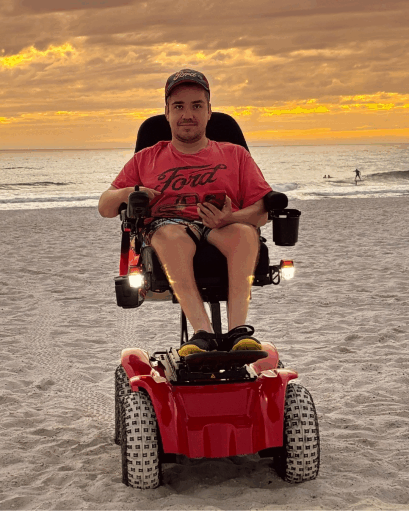 A man in a red motorized wheelchair sits on a sandy beach at sunset, wearing a red T-shirt, shorts, and a cap, holding a phone. The ocean and a few distant people are visible in the background.