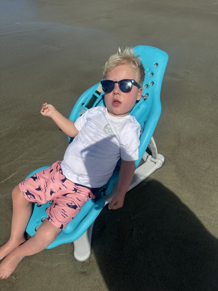 A young boy wearing sunglasses, a white shirt, and pink swim trunks with blue sharks relaxes on a blue beach chair on wet sand, enjoying a sunny day at the beach—this is just another joyful chapter in Myles' Story.