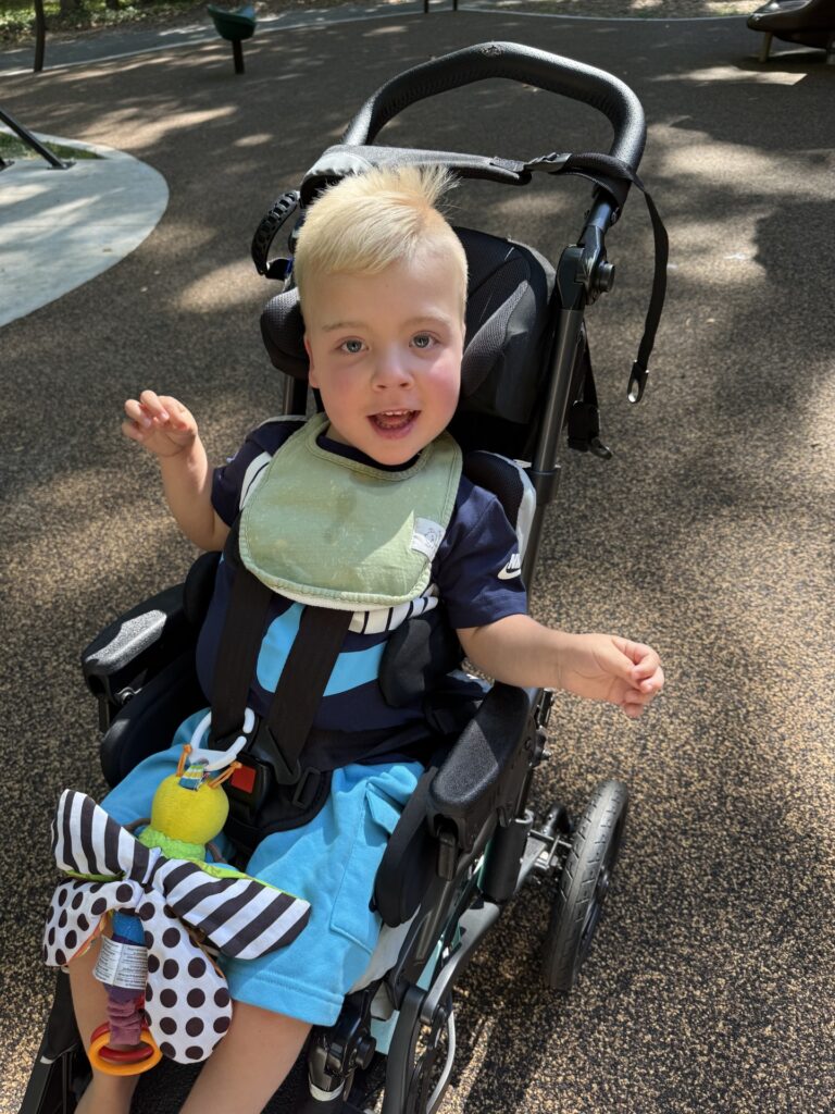 A smiling young child with blonde hair sits in a stroller at a playground, wearing a bib and holding a toy with a striped bow and pacifier attached—this joyful moment is part of Myles' Story as sunlight filters through the trees.