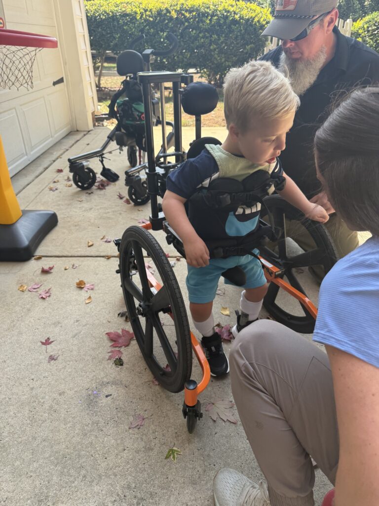 A young boy in a standing wheelchair is supported by an adult. Nearby, another adult and a second wheelchair are on a driveway scattered with autumn leaves, next to a toy basketball hoop and garage—capturing a moment from Myles' Story.