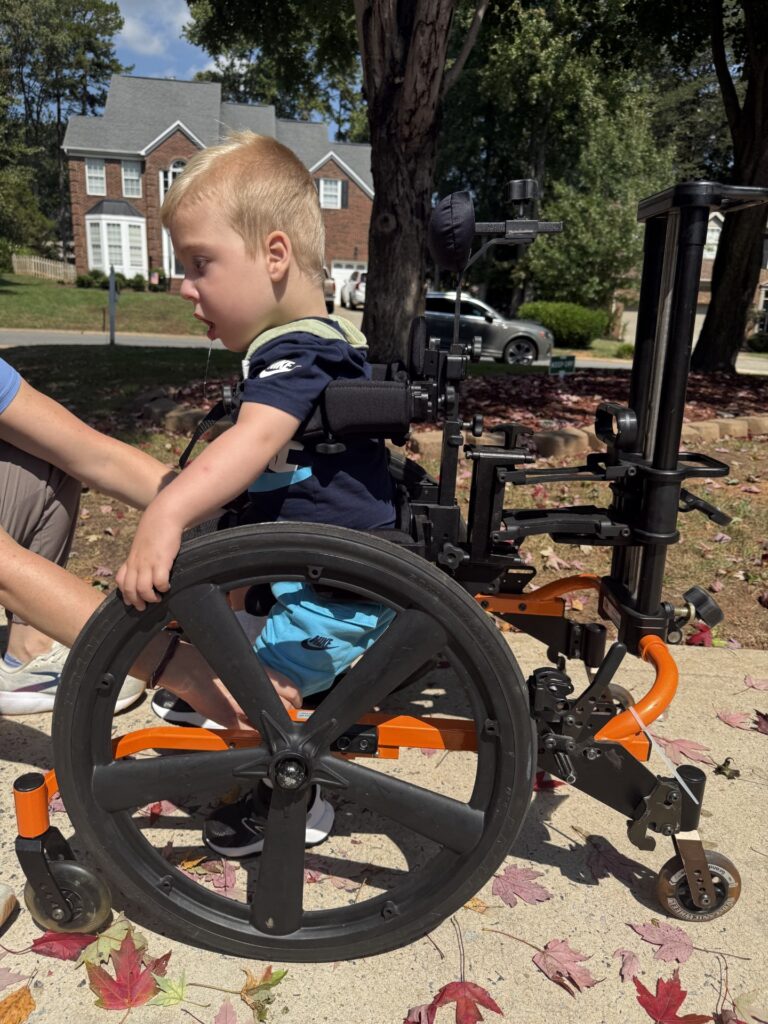 A young boy with light hair sits in a specialized wheelchair outdoors on a sunny day, while an adult assists him. Fallen red and brown leaves are scattered on the ground, setting the scene for Myles' Story among homes in the background.