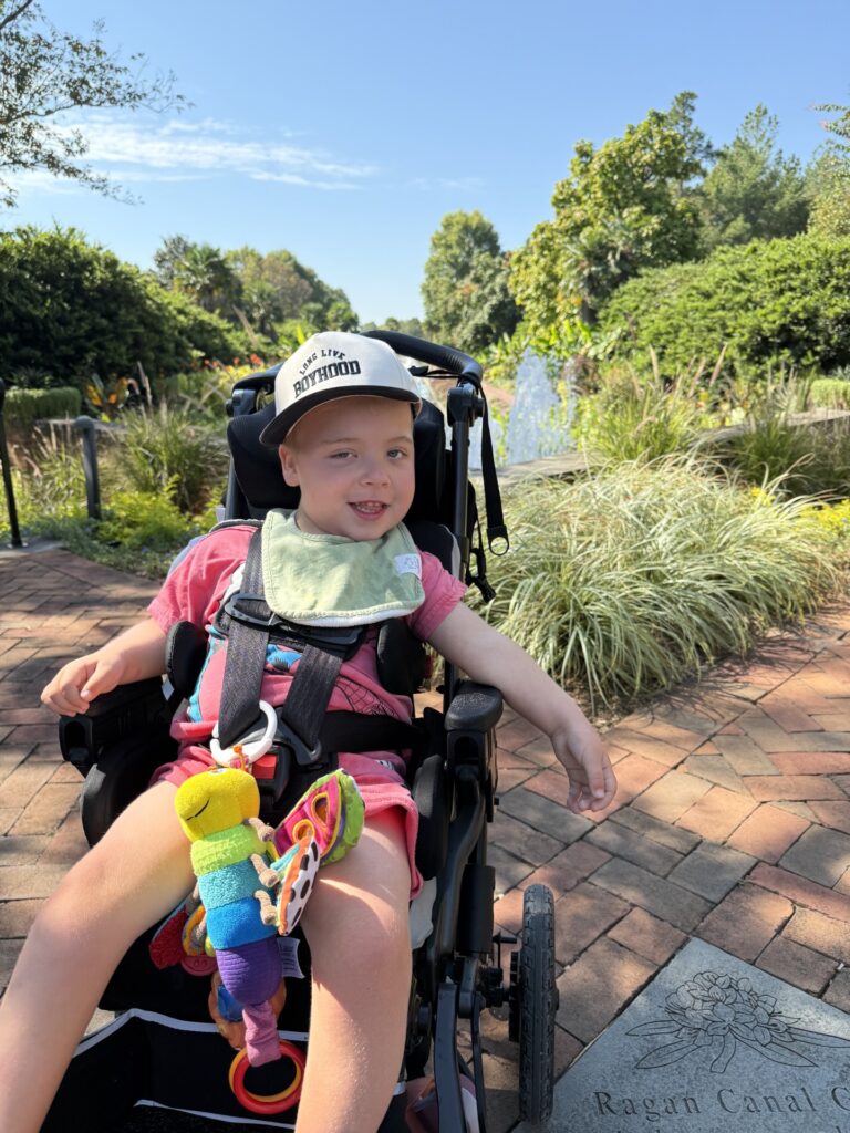 A smiling child in a stroller wears a white cap and pink outfit, holding a colorful toy. Myles’ Story unfolds outdoors on a sunny day, with greenery, a brick path, and a fountain in the background.