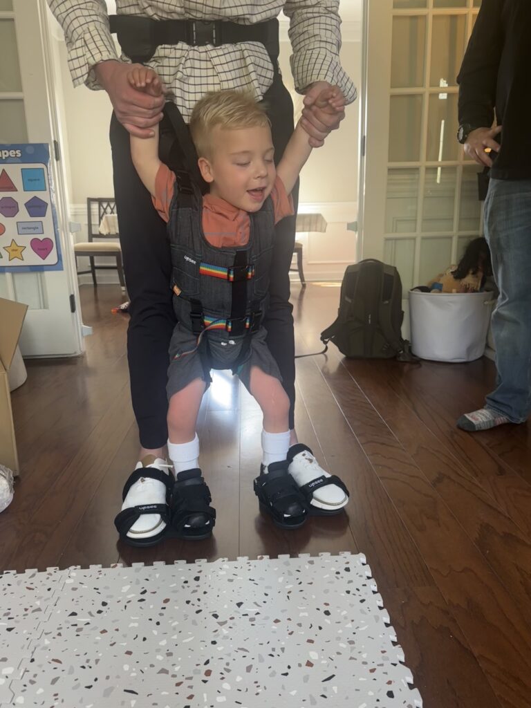 A smiling young boy wearing supportive braces and sandals is held up by an adult standing behind him, helping him stand and walk inside a sunlit room—an inspiring moment in Myles' Story.