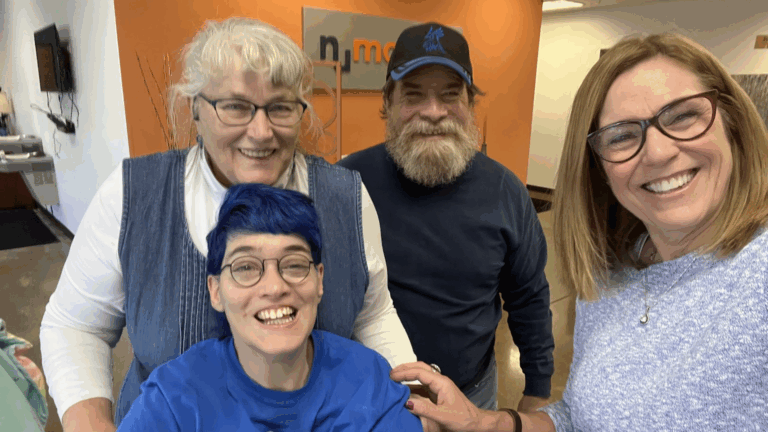Four adults smiling for a group selfie indoors; capturing Michele's Story, one has blue hair and glasses, another wears a white shirt and has gray hair. A man with a beard sports a blue cap while a woman with glasses stands on the right by the orange wall.
