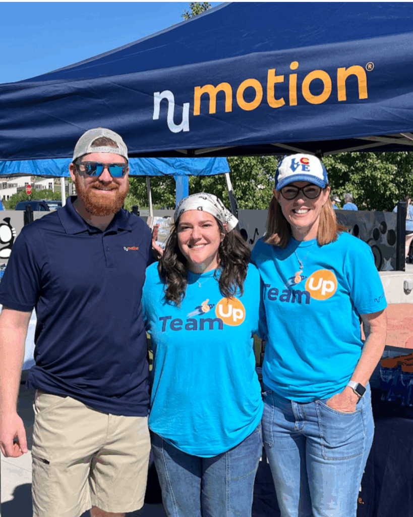 Three people stand smiling under a blue tent with the nu motion logo. Two wear matching blue Team Up shirts and sunglasses; the third, featured in Michele's Story, wears a navy shirt, khaki shorts, and a cap. It is sunny and bright outside.