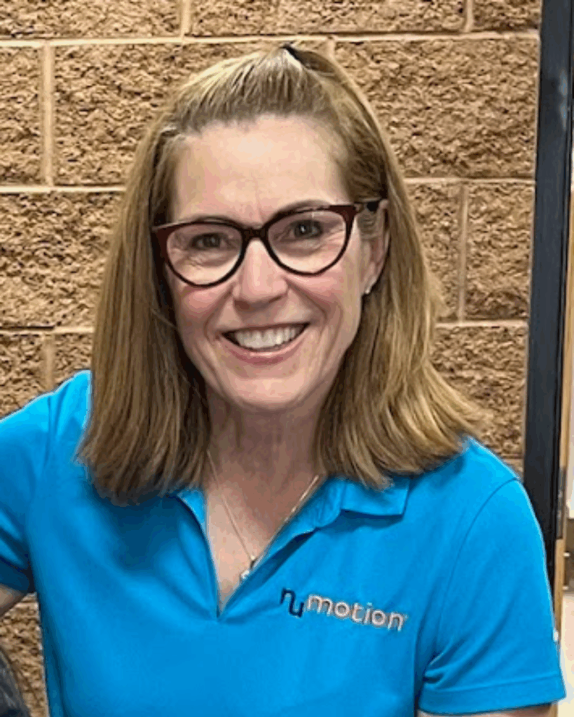 A woman with shoulder-length light brown hair and glasses smiles at the camera. She is wearing a blue polo shirt with the humotion logo, standing in front of a brick wall, capturing a moment from Michele's Story.