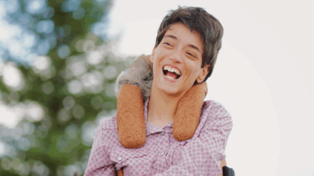 A young person with short hair smiles joyfully outdoors, wearing a light pink patterned shirt. A plush animal is draped around their neck, and greenery is softly blurred in the background—capturing a moment from Kylie’s Story.