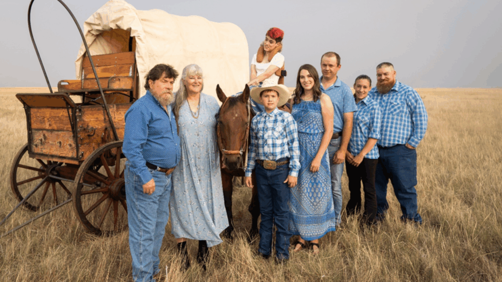 Eight people, including children and adults, pose outdoors in front of a covered wagon on a grassy plain. Dressed in blue western-style clothing, they stand beside a brown horse, bringing Kylie's Story to life on the frontier.