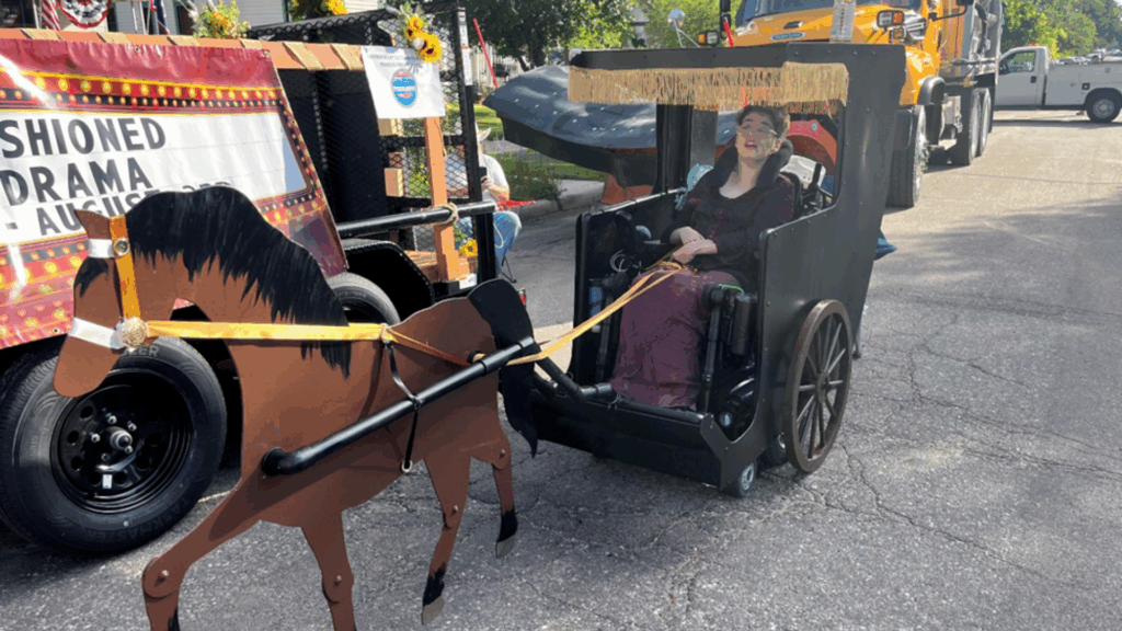 A smiling person in a wheelchair dressed as a carriage, holding reins attached to a cardboard horse, participates in the parade—bringing Kylie's Story to life amid brightly decorated floats and vehicles in the background.