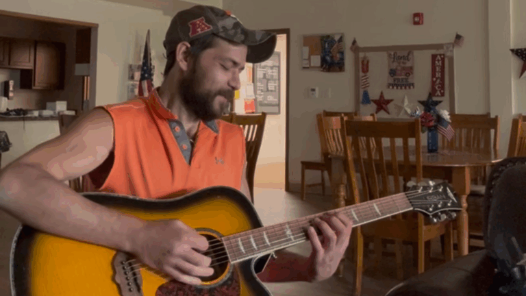 A man in an orange sleeveless shirt and camouflage cap plays an acoustic guitar in a room decorated with patriotic American items and wooden chairs and tables, capturing the spirit of Robert's Story.
