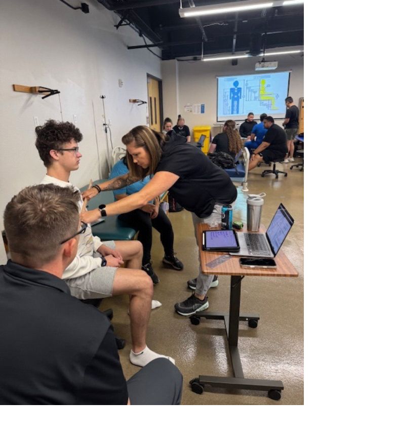 A woman checks a young mans arm with a device in a classroom setting, while others observe. A tablet and laptop are on a small table in the foreground. People are seated and engaged in the background.