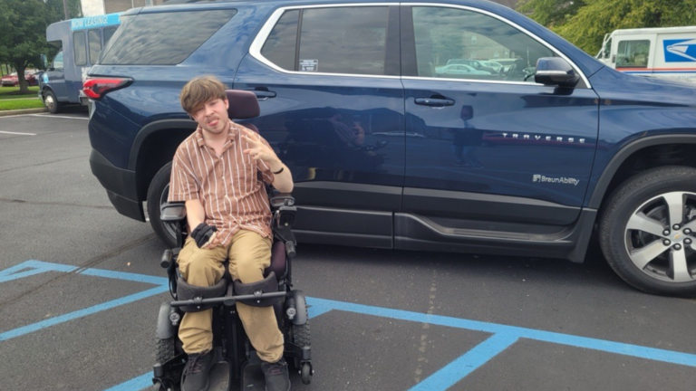 A man in a wheelchair poses and smiles in a parking lot next to a dark blue Chevrolet Traverse SUV parked in an accessible space, capturing a proud moment from Dillion's Journey. Trees and other vehicles are visible in the background.