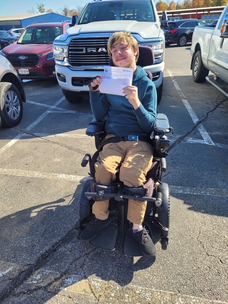 A person in a motorized wheelchair smiles and holds up a document in a parking lot, with several cars—including a white truck—visible behind them, capturing a joyful moment from Dillion's Journey.