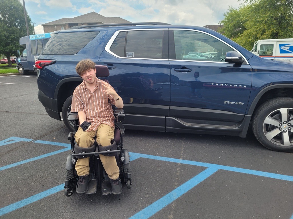 A young man in a powered wheelchair sits in a handicapped parking space next to a blue Chevrolet Traverse SUV with SmartAbility branding, highlighting Dillion's Journey. The weather is partly cloudy, with greenery and buildings in the background.