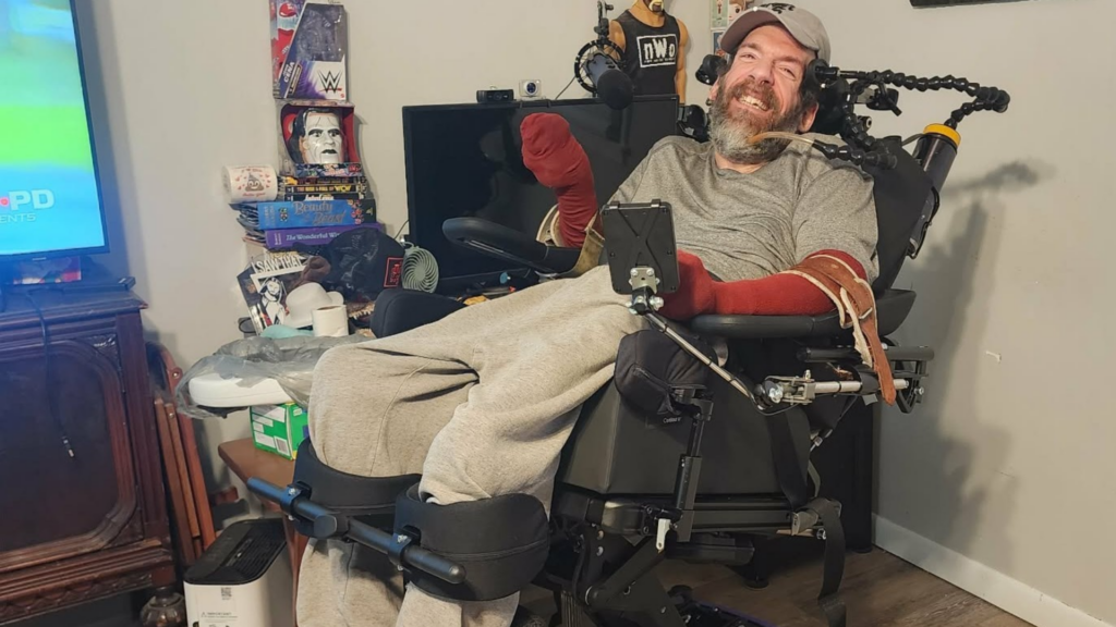 A man in a wheelchair smiles at the camera in a cozy room with wrestling memorabilia, books, and a TV. He wears a gray shirt, sweatpants, and has arm and leg supports. A tablet is mounted on his wheelchair.