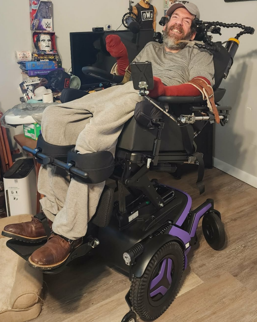 A smiling man with a gray beard sits in a motorized wheelchair with purple accents. He wears a gray shirt, sweatpants, brown shoes, and arm supports. Behind him are shelves with books, electronics, and collectibles.