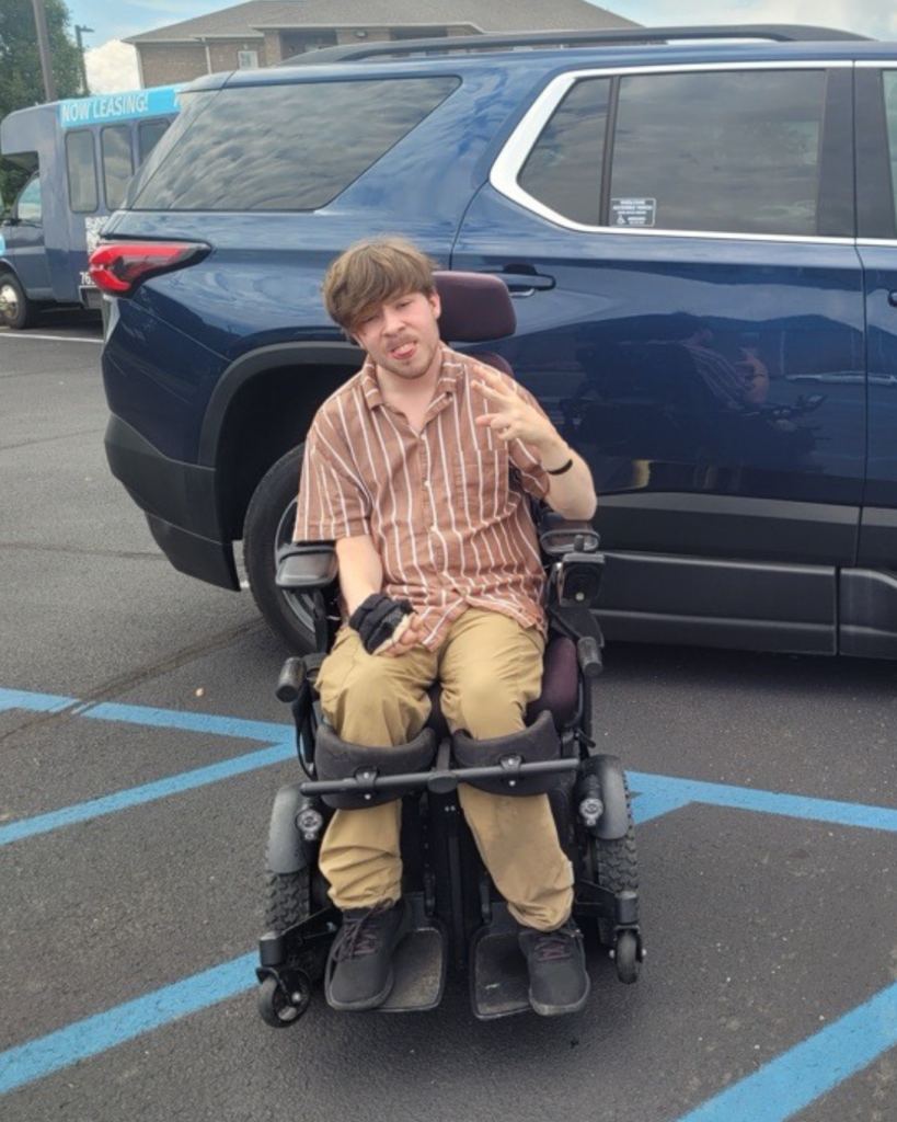 A young man in a wheelchair sits in a parking lot, wearing a striped shirt and khaki pants, making a peace sign with his right hand. A blue SUV is parked behind him, and blue handicap parking lines are visible.