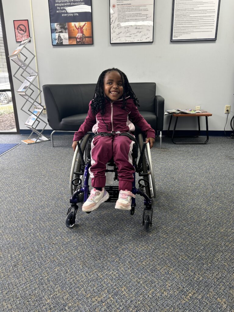 A young child smiles while sitting in a wheelchair, wearing a matching maroon tracksuit and white sneakers, inside a waiting room with a black couch, posters on the wall, and a magazine rack—capturing the warmth of Jaliece’s Story.