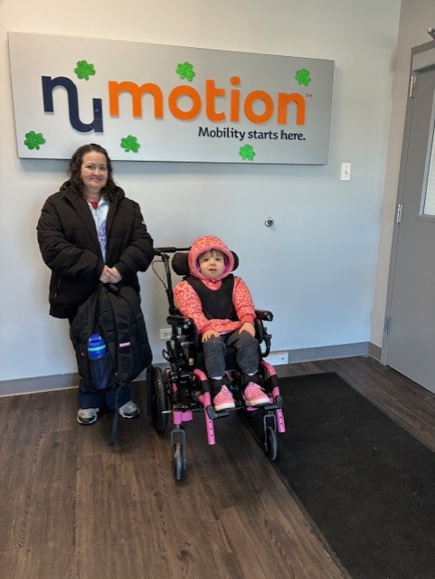 Two people are in an office lobby. A child in a pink jacket sits in a wheelchair, and an adult stands beside her, both near a sign that reads Numotion: Mobility starts here—this is part of Violet's Story.