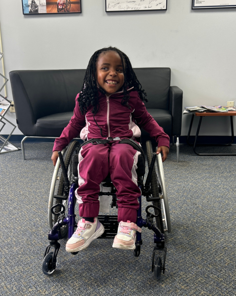 A young girl in a maroon tracksuit sits and smiles in a wheelchair in a waiting area with a black couch, a small table with magazines, and framed photos on the wall behind her.