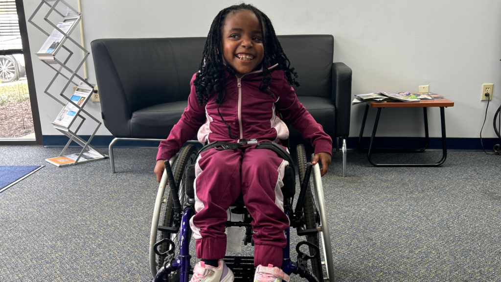 Jaliece, a young girl with long braids, wearing a maroon tracksuit and pink sneakers, smiles while sitting in a wheelchair inside an office offering a one-stop service, with a gray couch and small table in the background.